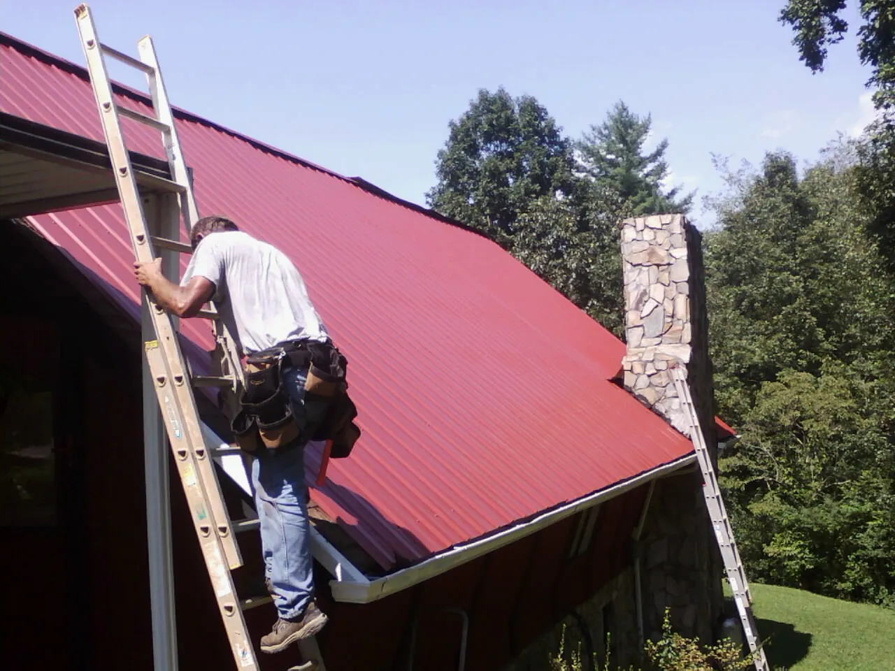 Roofer on a ladder beside a red metal roof and stone chimney — Soffit & Fascia Repair by Roofing Brothers in East Rochester